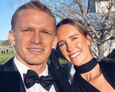cody simpson and emma mckeon pose against a blue sky. Cody wears a black suit and bowtie, emma wears a black dress