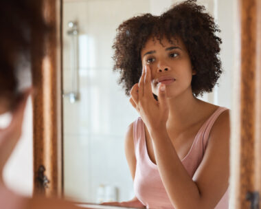 Young African woman putting moisturizer on her face while getting ready in her bathroom in the morning