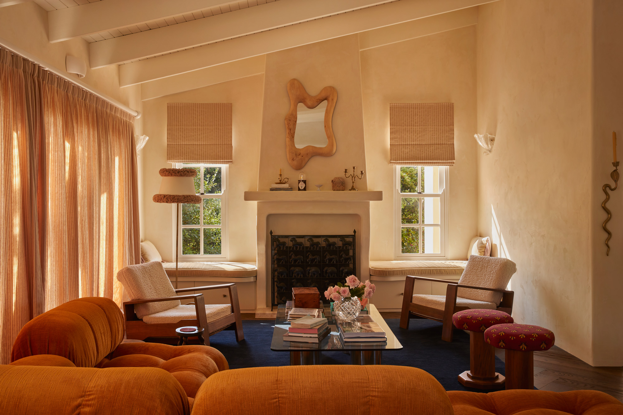 the loung room inside the home of chloe tozer, featuring a blue rug, sand-coloured walls, two arm chairs and an orange sofa set around a glass coffee table and a fireplace