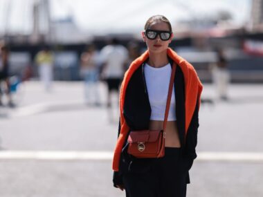 woman stands in street with orange jumper over her shoulders and a crossbody bag