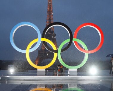 a shot of the olympic rings, lit up at night, with the Eiffel Tower as a shadow in the background, taken ahead of the 2024 Olympics in Paris
