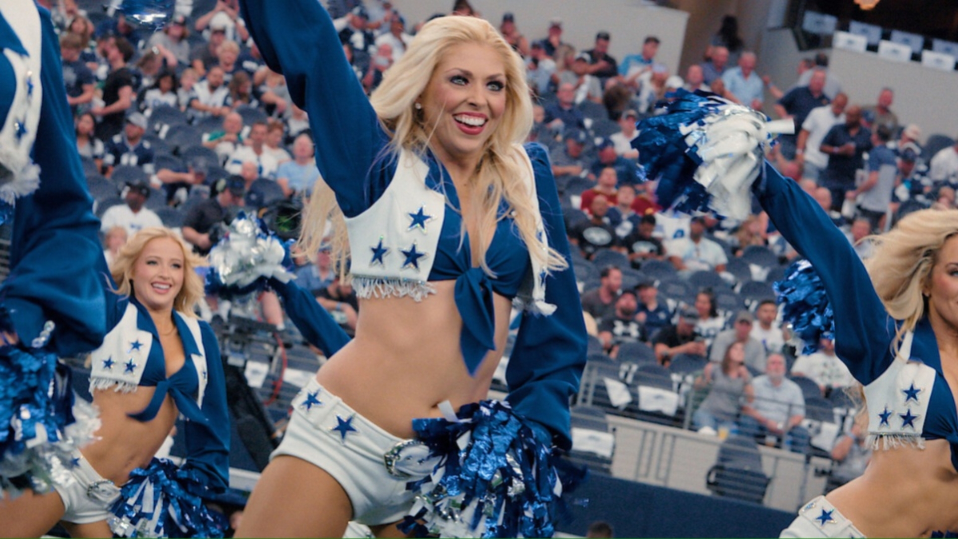 A blonde Dall cowboys cheerleader wears white shorts and a white and blue top, holding pom pons with a hand above her head and one on her hip