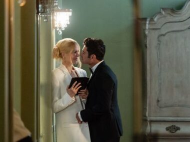 A man and woman, dressed formally, share a tender moment in a room with an elegant wardrobe and chandelier; the woman holds a book.