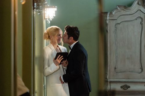 A man and woman, dressed formally, share a tender moment in a room with an elegant wardrobe and chandelier; the woman holds a book.