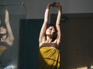 A woman with curly hair stretches post shower, wrapped in a yellow towel with a smile on her face.