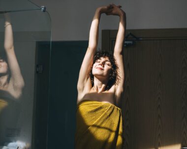 A woman with curly hair stretches post shower, wrapped in a yellow towel with a smile on her face.