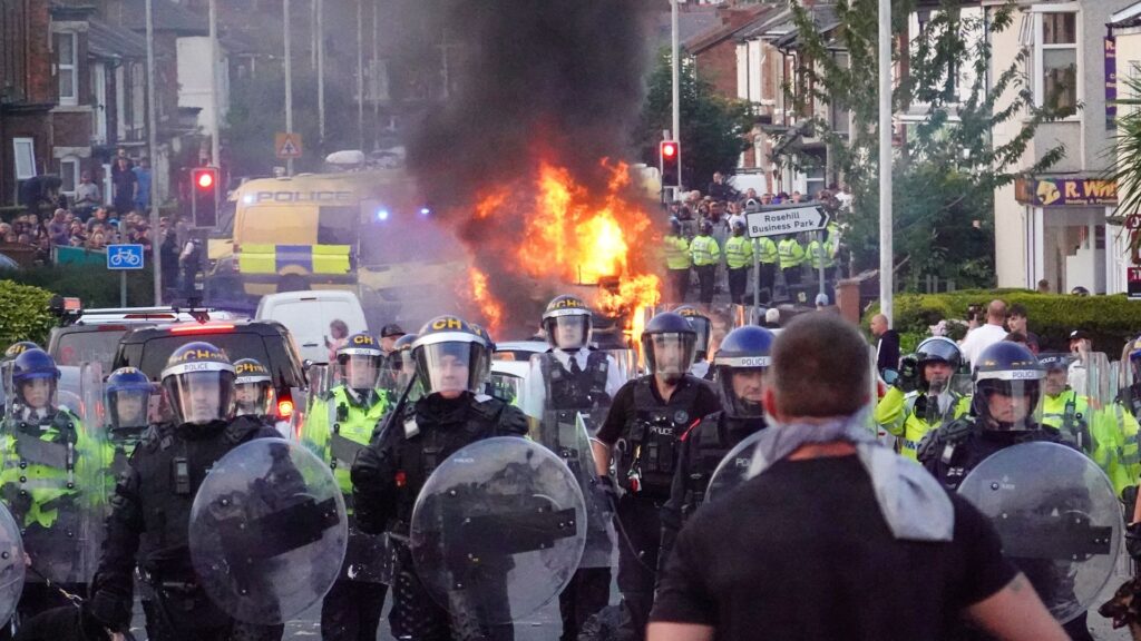 Riot police hold back protesters near a burning police vehicle after disorder broke out on July 30, 2024 in Southport, England. Rumours about the identity of the 17-year-old suspect in yesterday's deadly stabbing attack here have sparked a violent protest. According to authorities and media reports, the suspect was born in Cardiff to Rwandan parents, but the person cannot be named due to his age. A false report had circulated online that the suspect was a recent immigrant who crossed the English Channel last week and was "on an MI6 watchlist."