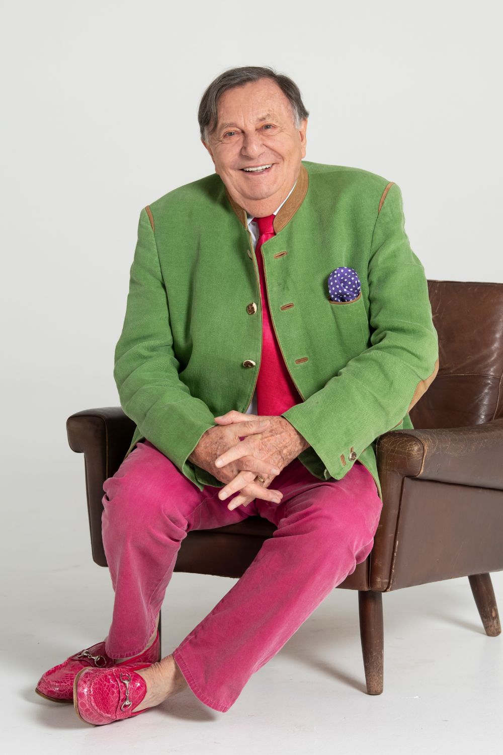 barry-humphries-sitting-in-leather-chair-in-green-blazer-and-pink-trousers-smiling