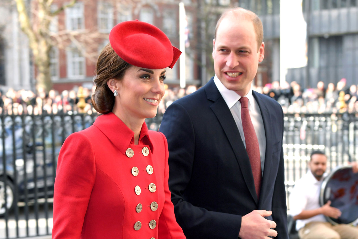 A woman in a red coat and matching hat stands beside a man in a suit. They are both smiling.