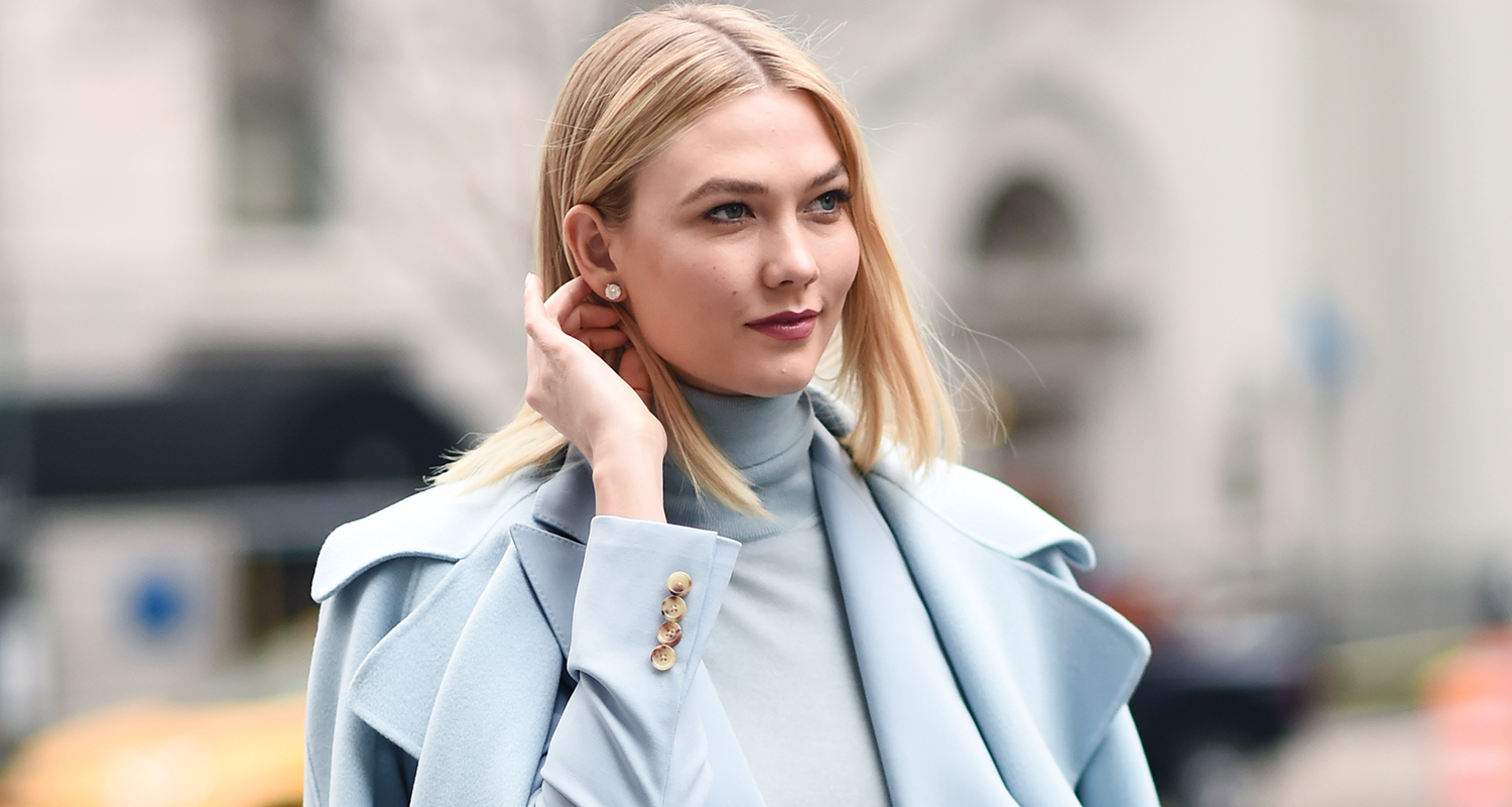 Woman with blonde hair in a light blue coat and turtleneck, outside in an urban setting.